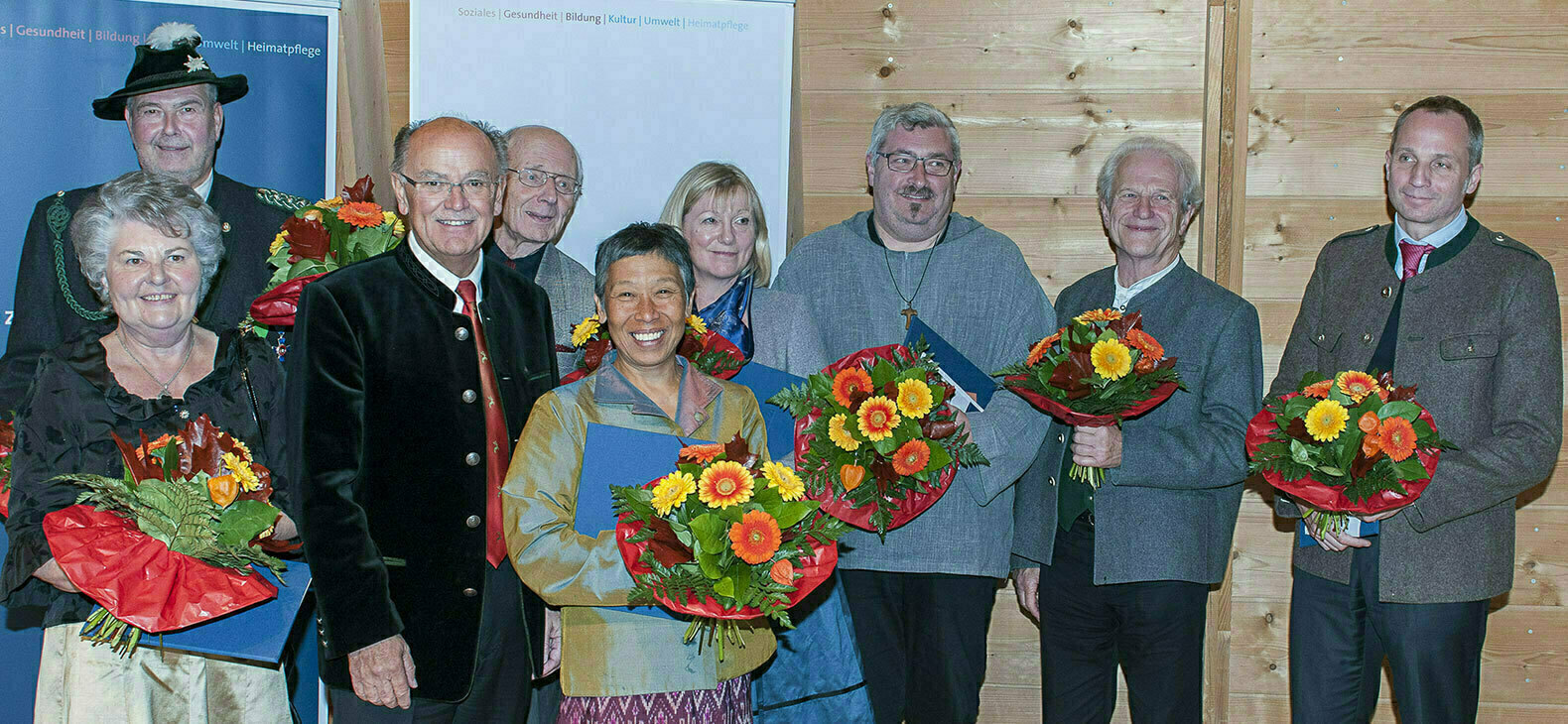 Gruppenfoto mit neun Personen vor einer Holzwand. Dritter von links ist Bezirkstagspräsident Josef Mederer. Ausser ihm halten alle einen Blumenstrauß in der Hand.