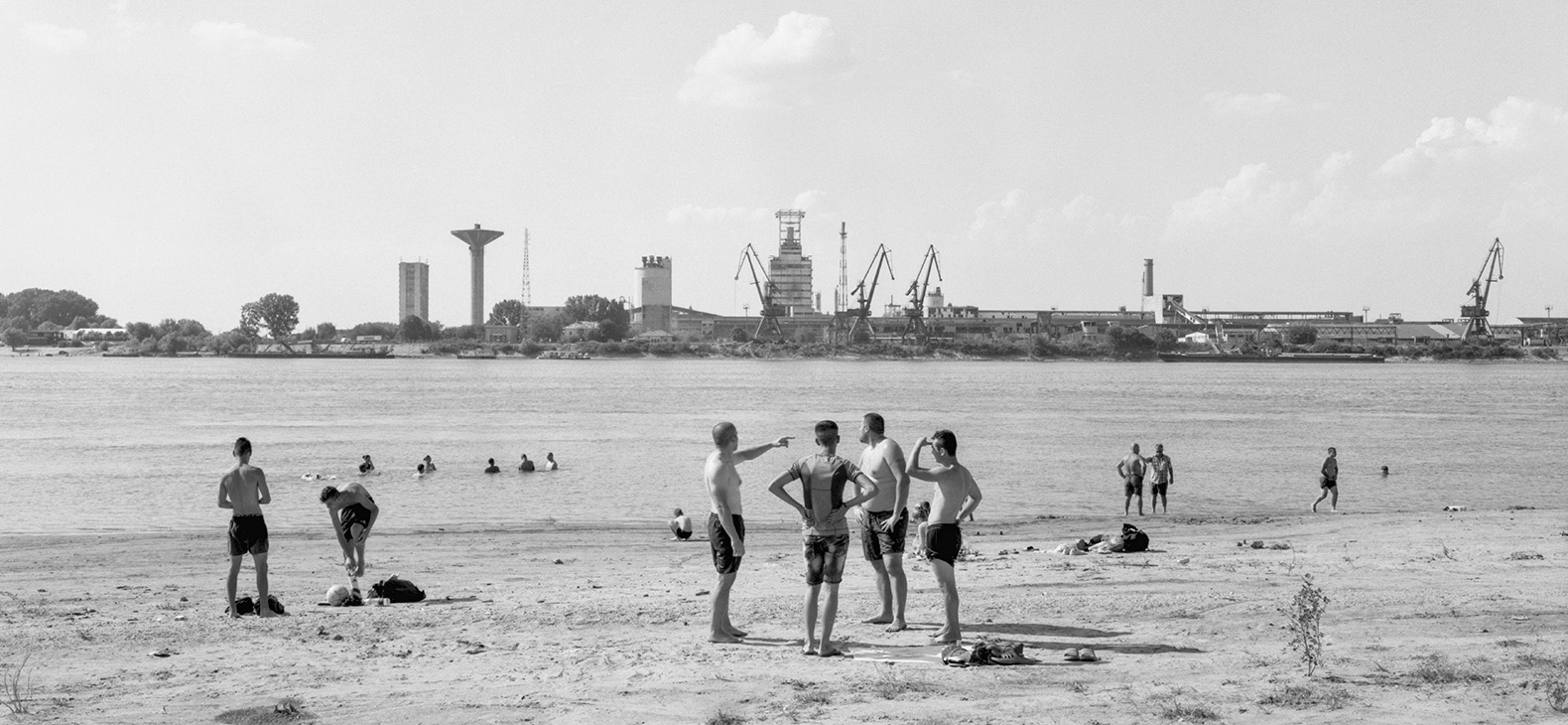 Ute Mahler and Werner Mahler: Danube #155, 2019, detail; silver gelatin print, 90 x 115 cm / b/w photo: group of people in the foreground on the bank of a large river, buildings and industrial plants in the background. 
