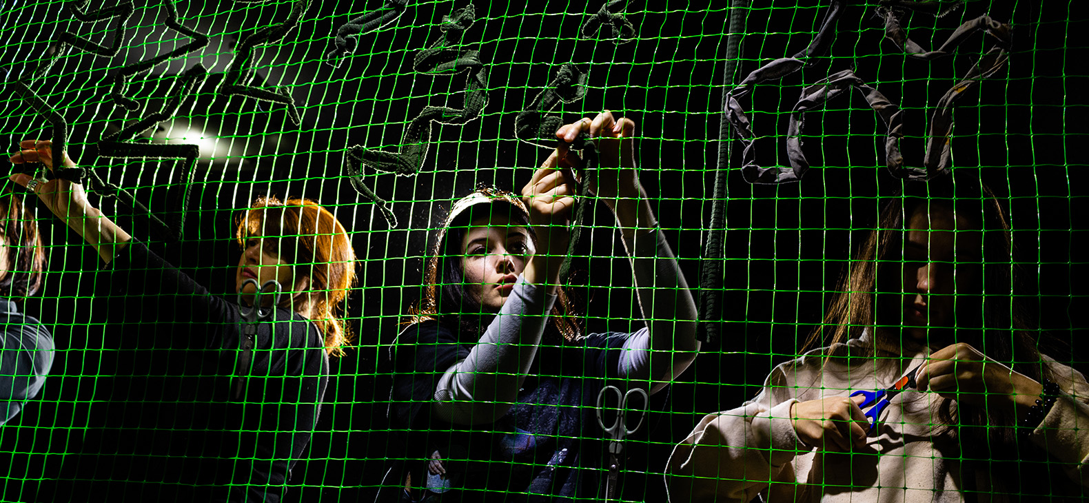 The Thin Line / Der schmale Grat - Ausstellung mit ukrainischen Fotografien; Bild: Freiwillige beim Knüpfen von Tarnnetzen, junge Frauen im Scheinwerferlicht hinter einem grünen Netz; Fotografin: Kateryna Moskalyuk 