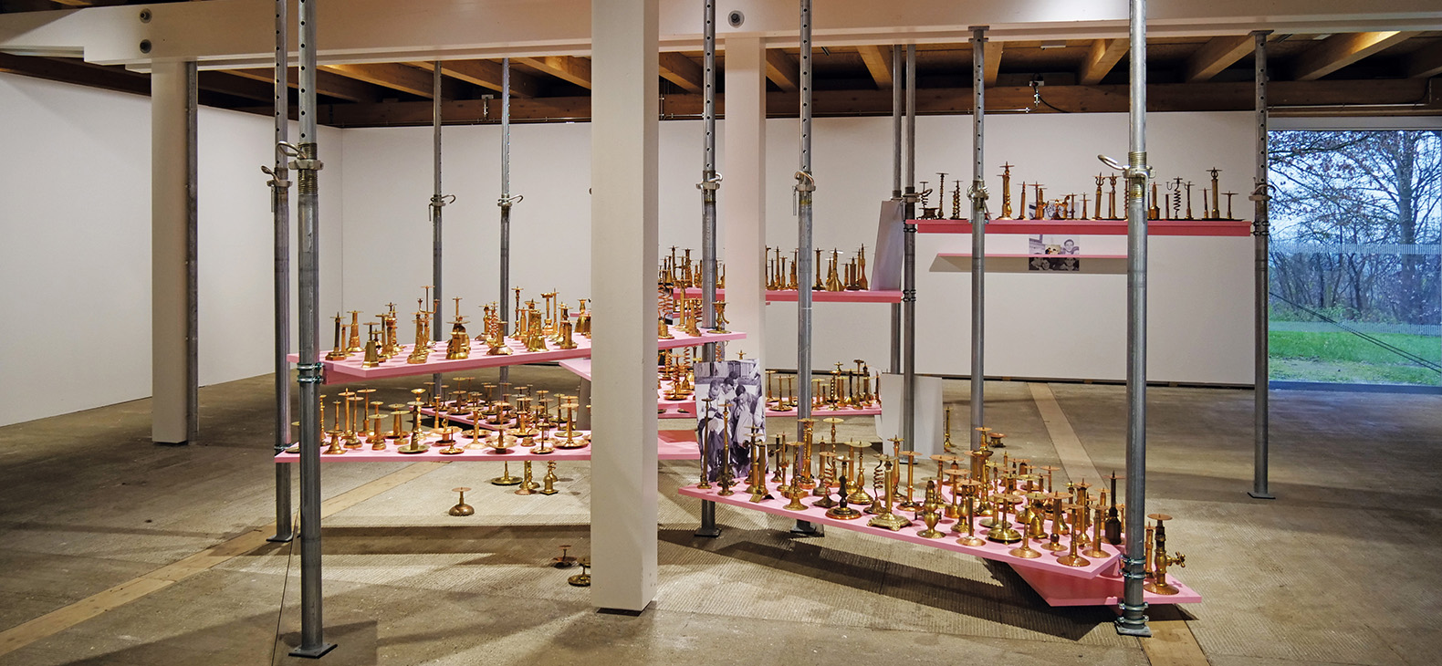 Blick in die Ausstellung; Installation "Die Aufstellung" von Rebekka Bauer mit mehreren hundert Metallobjekten auf pinkfarbenen Brettern zwischen Baustützen sowie freistehenden Fotoplatten