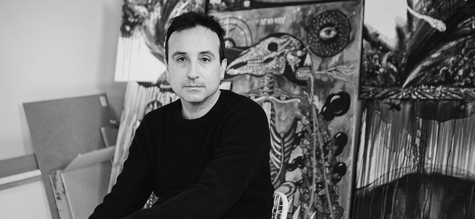 Black and white portrait of artist Olexa Mann in front of several of his works in a studio. 