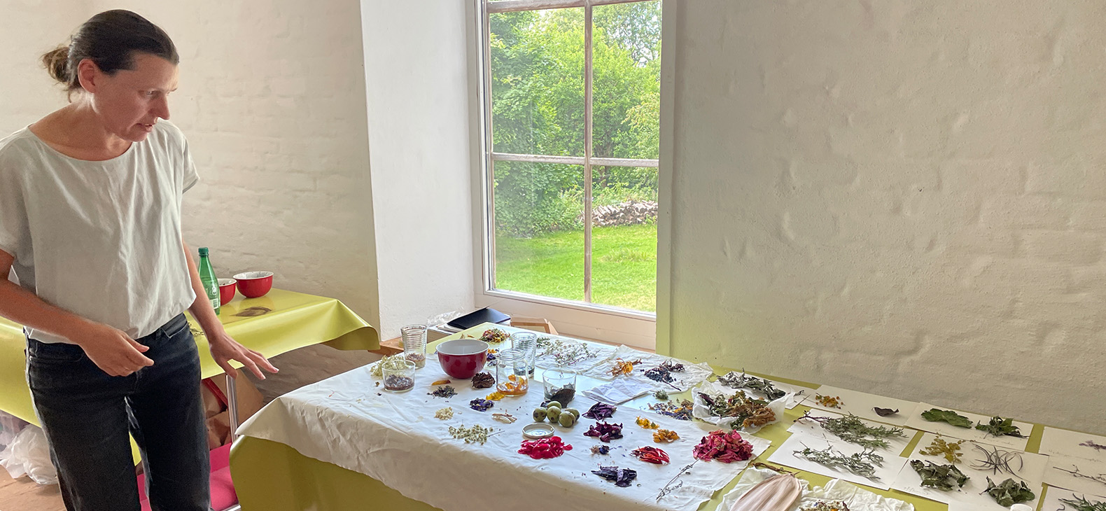 Artist Valerie Leray in her studio at the Schafhof - European Art Forum Upper Bavaria. She looks at her work material arranged on the table, which consists of plants, flowers and small fruits. In the background you can see through a window into the garden of the Schafhof.