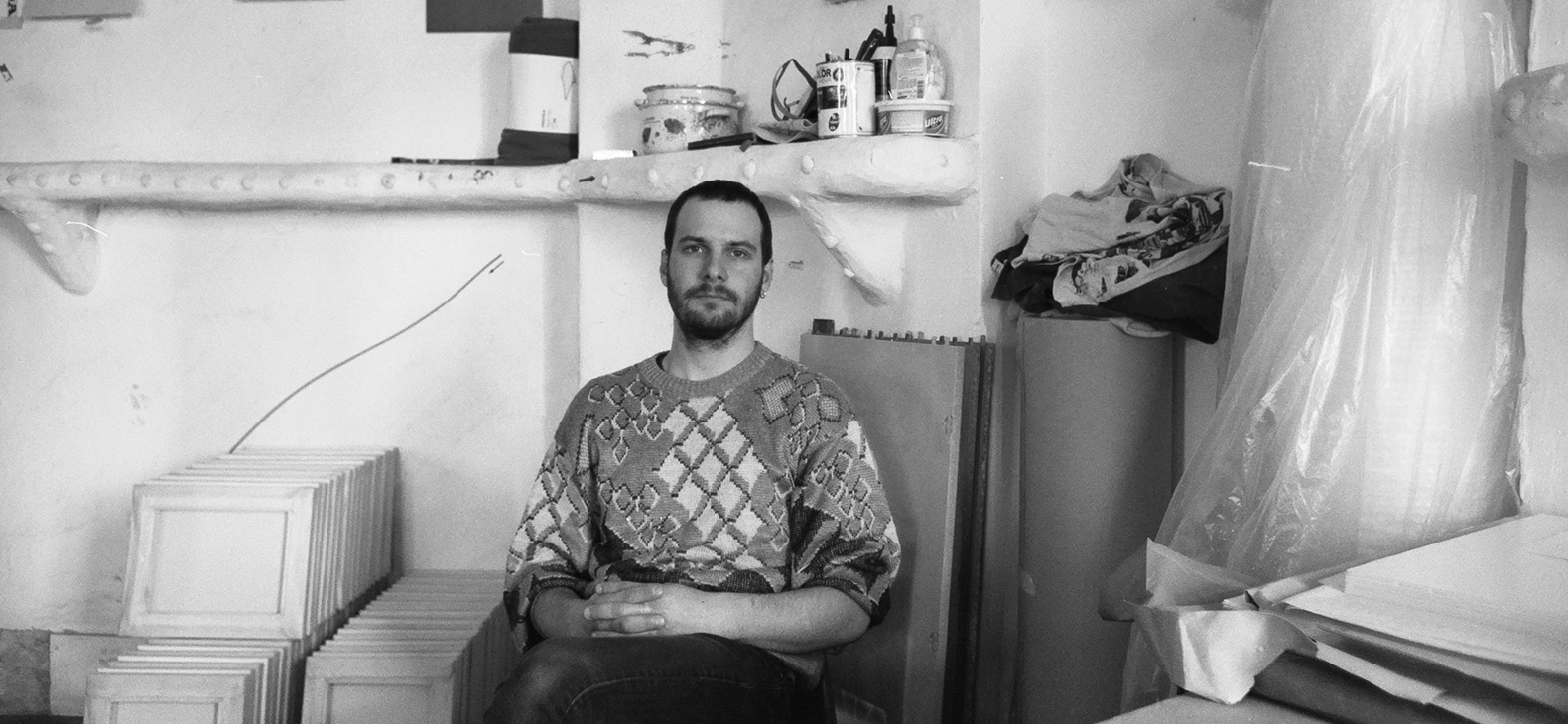 Schwarz-weiß Portrait des ungarischen Künstlers Ádám Varga sitzend in seinem Atelier.