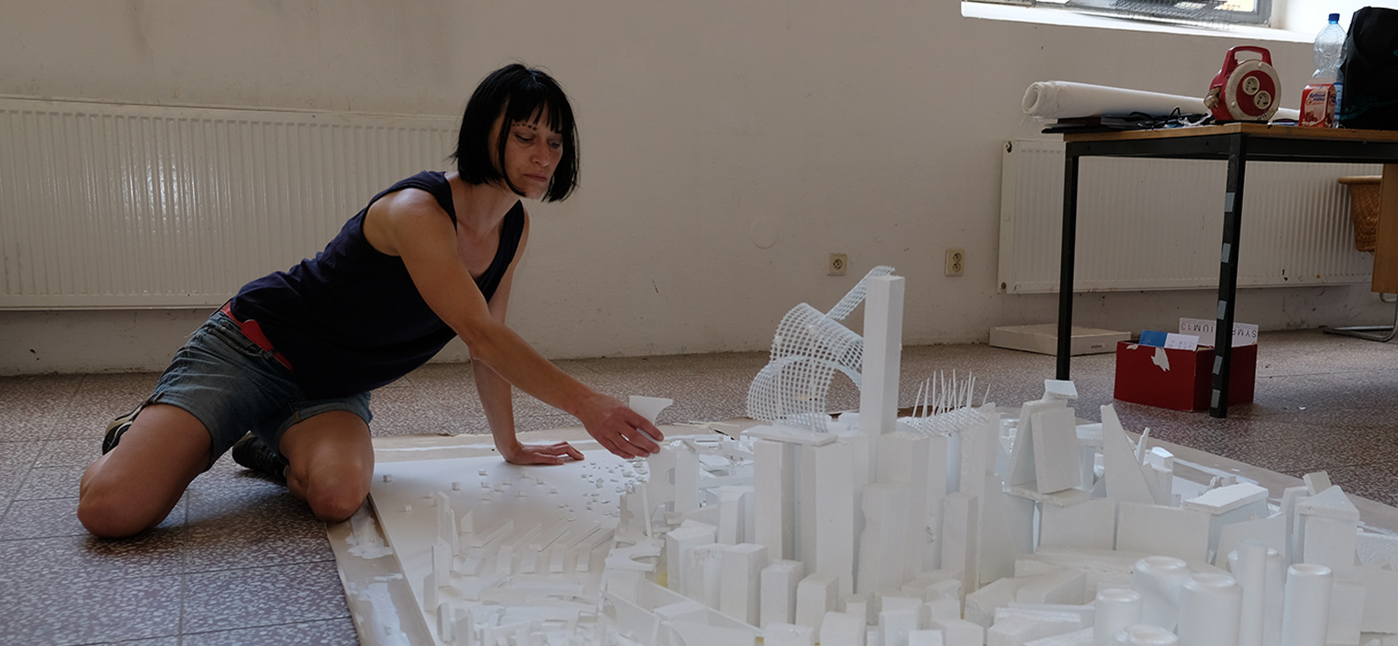 Schafhof Residency Programme; Artists 2023: Margit Greinöcker, image: artist kneeling on the floor over a work that presumably represents some kind of white model of a city