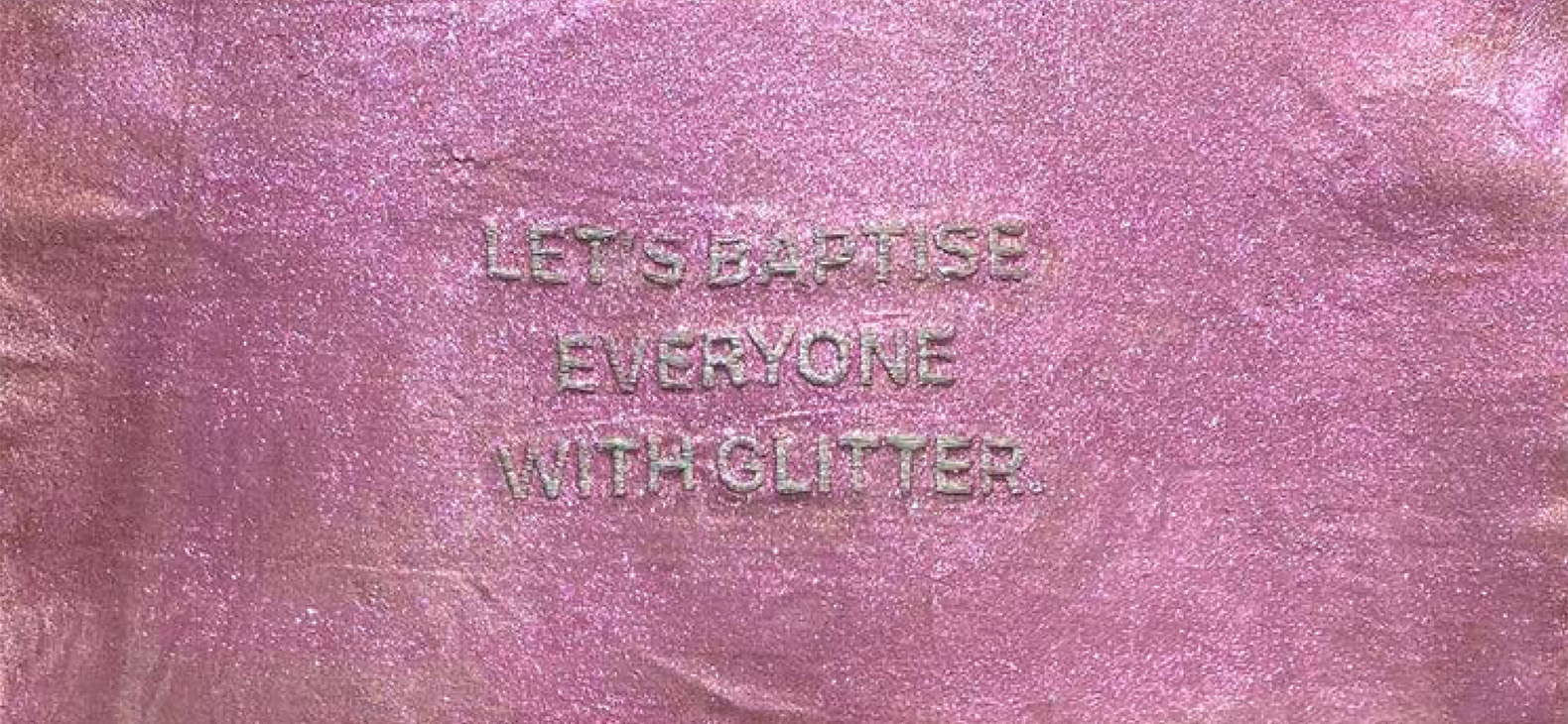 Das Bild zeigt ein Ausschnitt des Kunstwerks der Künstlerin Mariandrie Chrysostomou. Der Hintergrund ist Pink und glitzert. In der Mitte ist ein Schriftzug in glitzernder silberner Schrift. Der Schriftzug lautet "LET'S BAPTISE EVERYONE WITH GLITTER".
