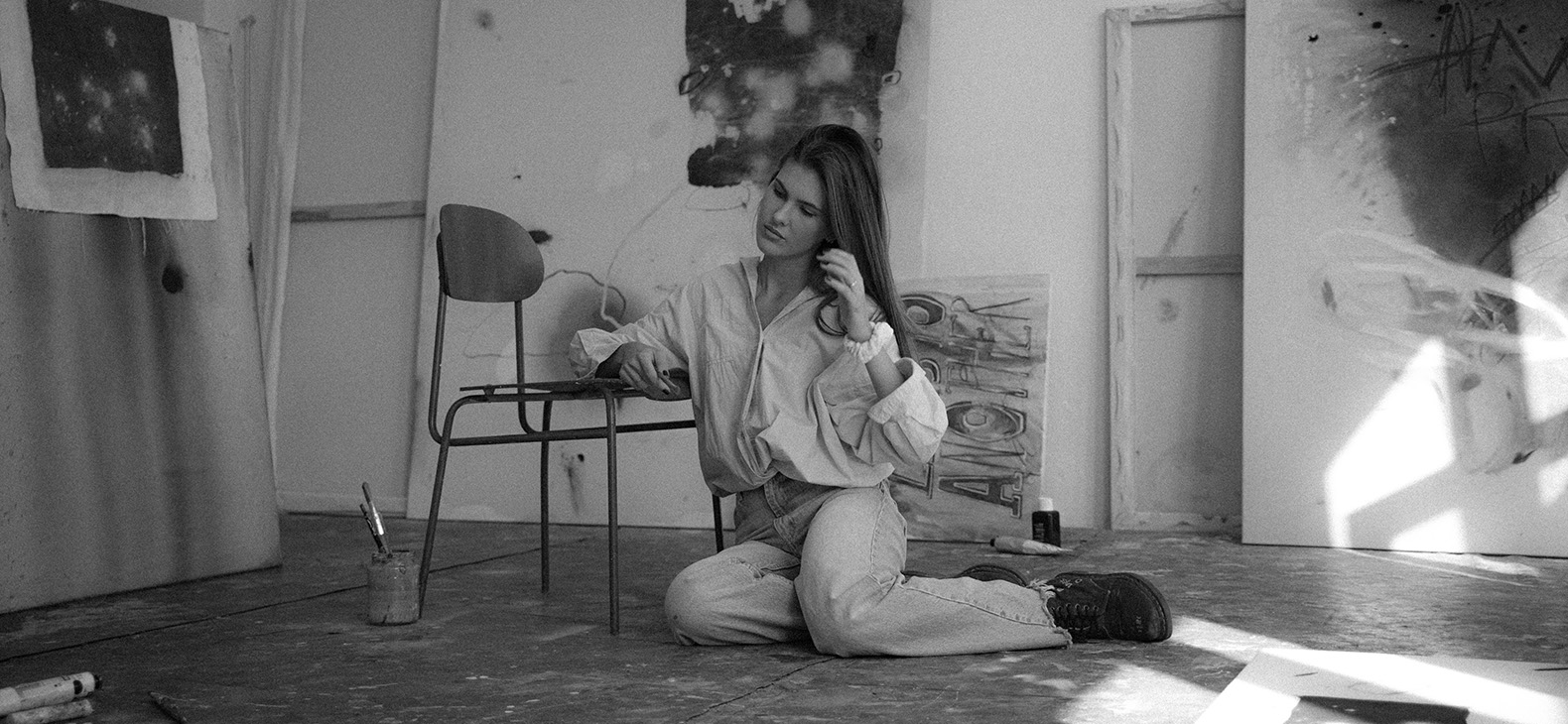 Rachel Jutka: Aufenthalt am Schafhof im Herbst 2023. Schwarz-weißes Bild der Künstlerin. Sie sitzt in ihrem Studio auf dem Boden und leht sich mit ihrem rechtem Arm auf einem Stuhl an, der neben ihr steht.
