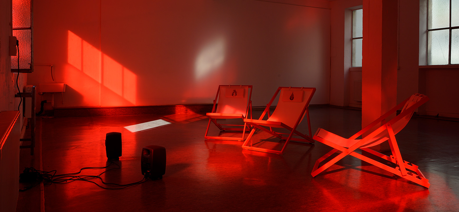Photo of the artwork "The Flamekeepers" by the artist Lilian Robl. It is a room installation from 2023. You can see a red room in which there are three deck chairs, each with a flame printed on its head pad. Two loudspeakers are set up on the floor in front of the chairs.