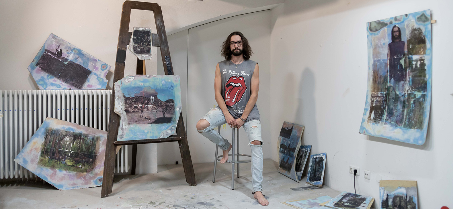 The artist Petr Kubac sits on a stool. His paintings, which he worked on during his stay at the Schafhof, can be seen in the room and on the walls.