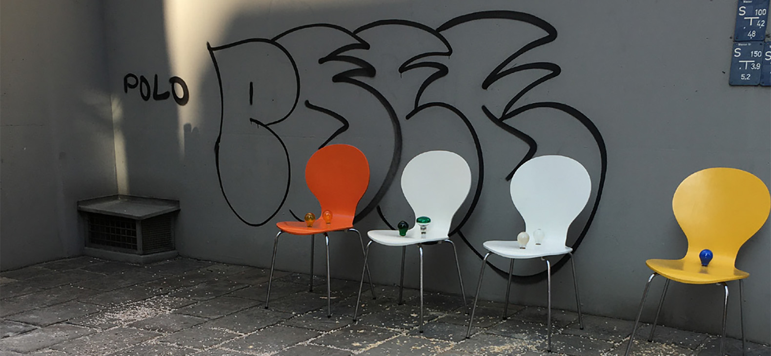 Mascha Illich: zero waste, Lockdown-Interventionen im öffentlichen Raum, 2021. In dem Bild sind vier Stühle zu sehen, die an einer grauen Wand stehen. Auf der Wand ist ein schwarzes Grafitti. Auf den Stühlen stehen 1-2 Glühbirnen.