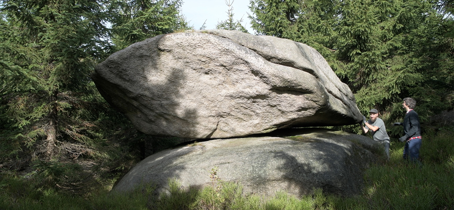 Metastabli: Auf dem Bild sieht man eine wäldliche Umgebung, in der Mitte des Bildes ist ein großer Stein zu sehen der auf einem anderen balanciert. Rechts der Steine sieht man die zwei Künstler wie sie den oberen Stein zum schaukeln bringen.