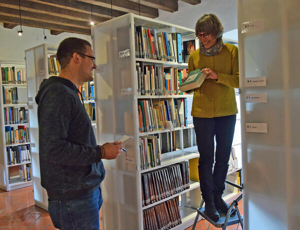 In einem Raum mit vielen Bücherregalen steht eine Frau auf einer Leiter. Sie hält ein Buch in der Hand, das sie einem Mann zeigt, der neben ihr steht.